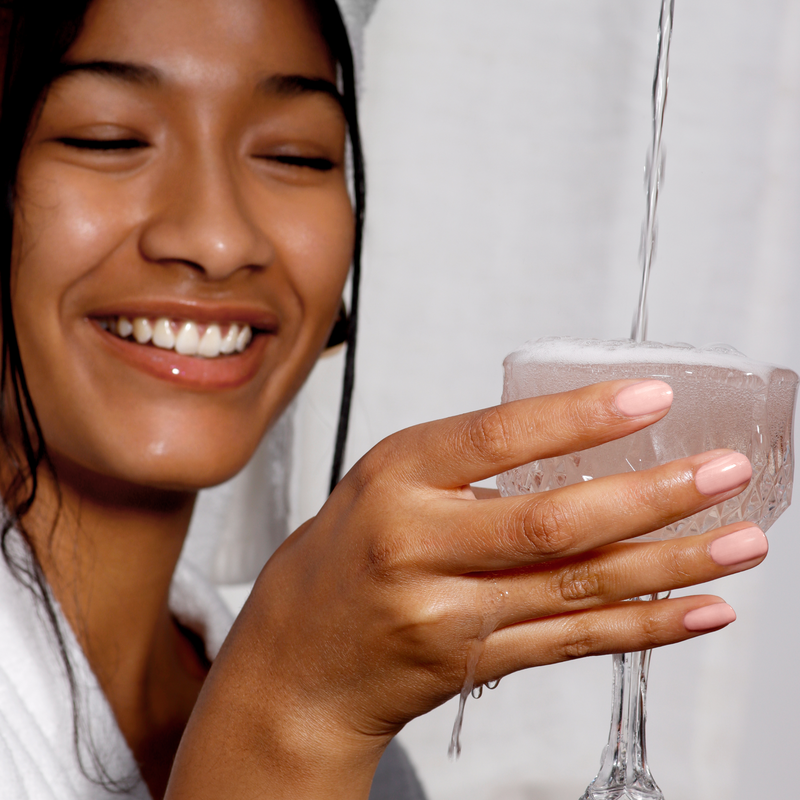 A person warmly smiles while holding a glass being filled with water. They're clad in a white robe against a softly blurred backdrop with pink beige hues, evoking the soothing ambiance of LONDONTOWN's Rose Latte.