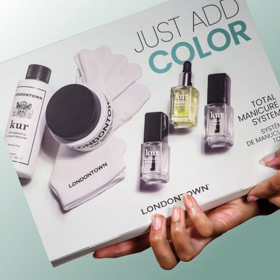 A person holds a boxed LONDONTOWN Just Add color manicure system, which includes nail care products, gloves, and bottles for an easy at-home manicure.
