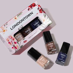 A box of LONDONTOWN Moonlit Botanica Mini lakur trio with three polishes—nude, glitter, and dark blue—arranged next to the box on a light surface.