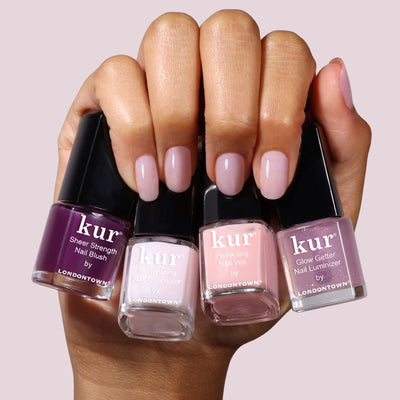 A hand with manicured nails holds four LONDONTOWN One-Step Hero Minis nail polishes in purple, pink, and nude shades against a light background.