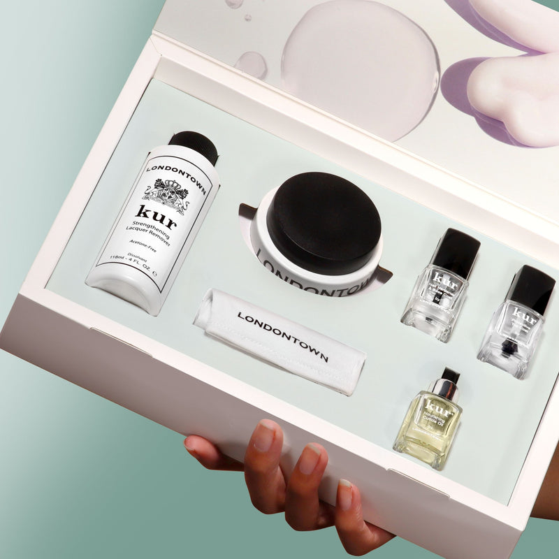 A hand holds an open box of LONDONTOWN's Just Add Color set, featuring nail care essentials like remover, treatment oils, a buffer, and nail polishes.
