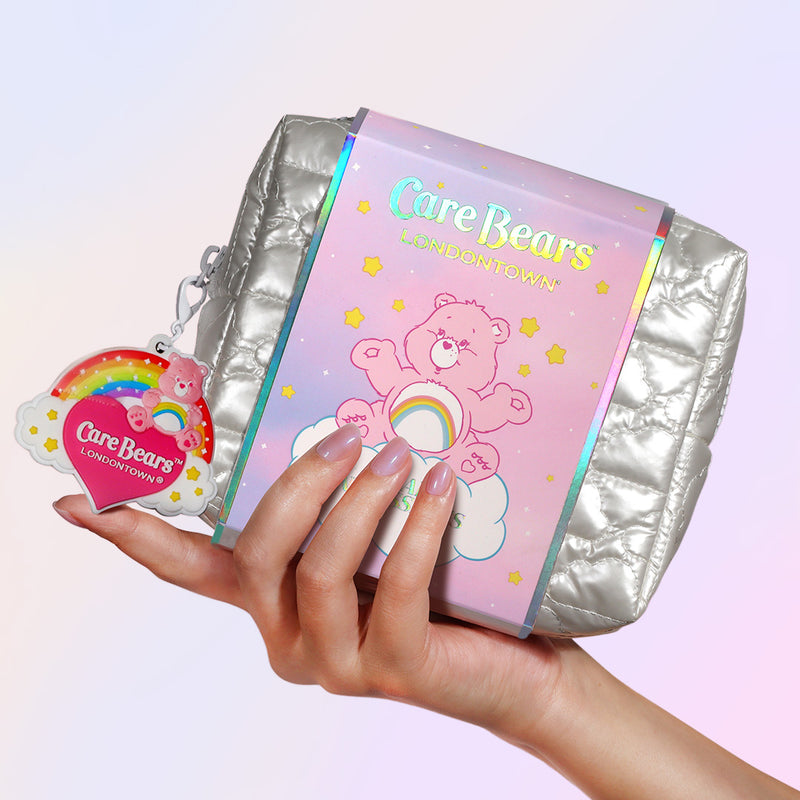 A hand holds the Bear Necessities Set by LONDONTOWN—a metallic silver pouch with Care Bears branding, a pink bear, rainbow design, and matching keychain tag—ideal for storing your essentials.