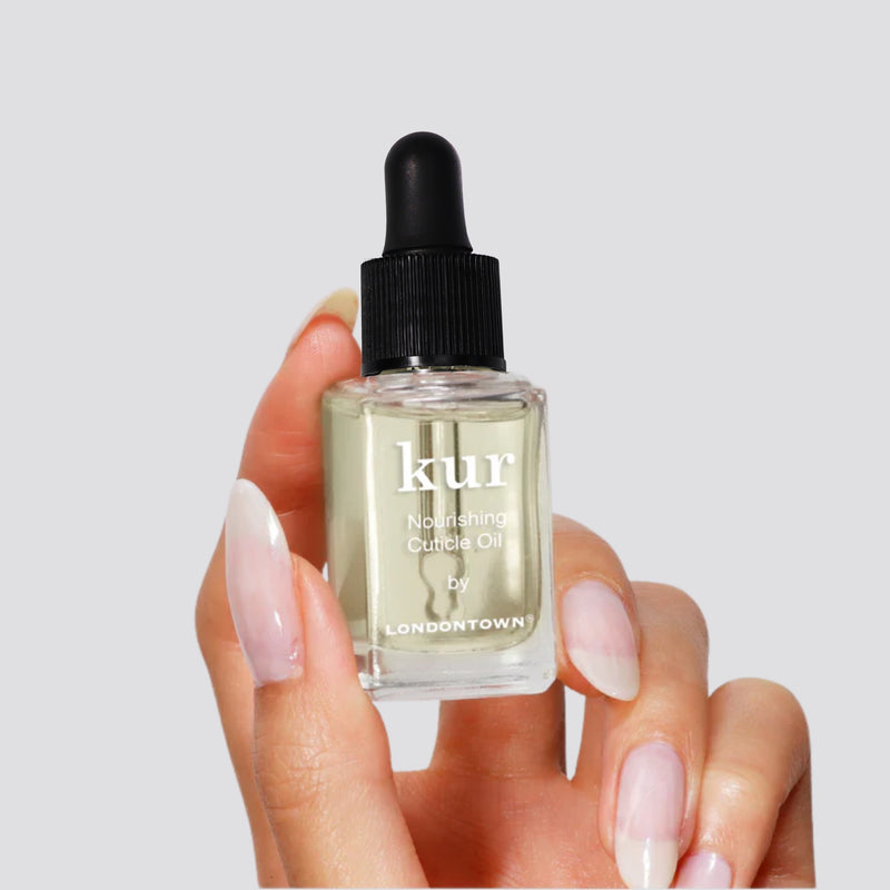 A manicured hand holds a small bottle of LONDONTOWN Nourishing Cuticle Oil with a black dropper cap—ideal for cuticle care and supporting nail health.
