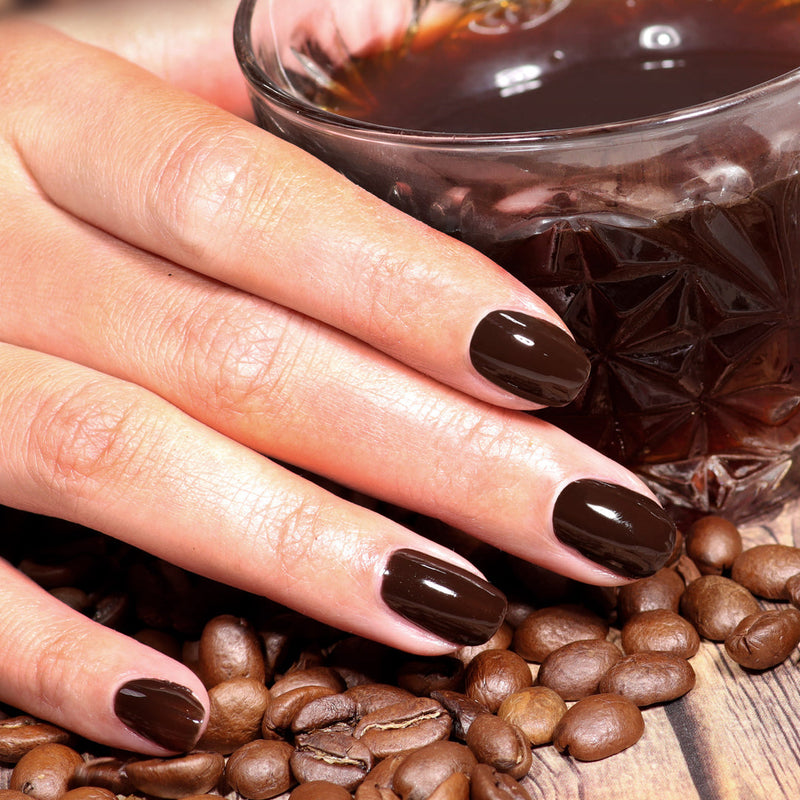 A hand with LONDONTOWN’s Midnight Mocha dark brown polished nails rests on coffee beans beside a glass of espresso.