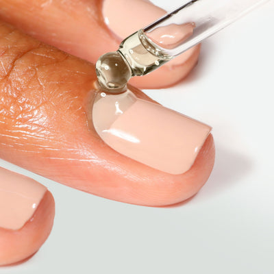 A close-up of a dropper applying LONDONTOWN's Just Add color clear oil onto a fingernail with a nude manicure.