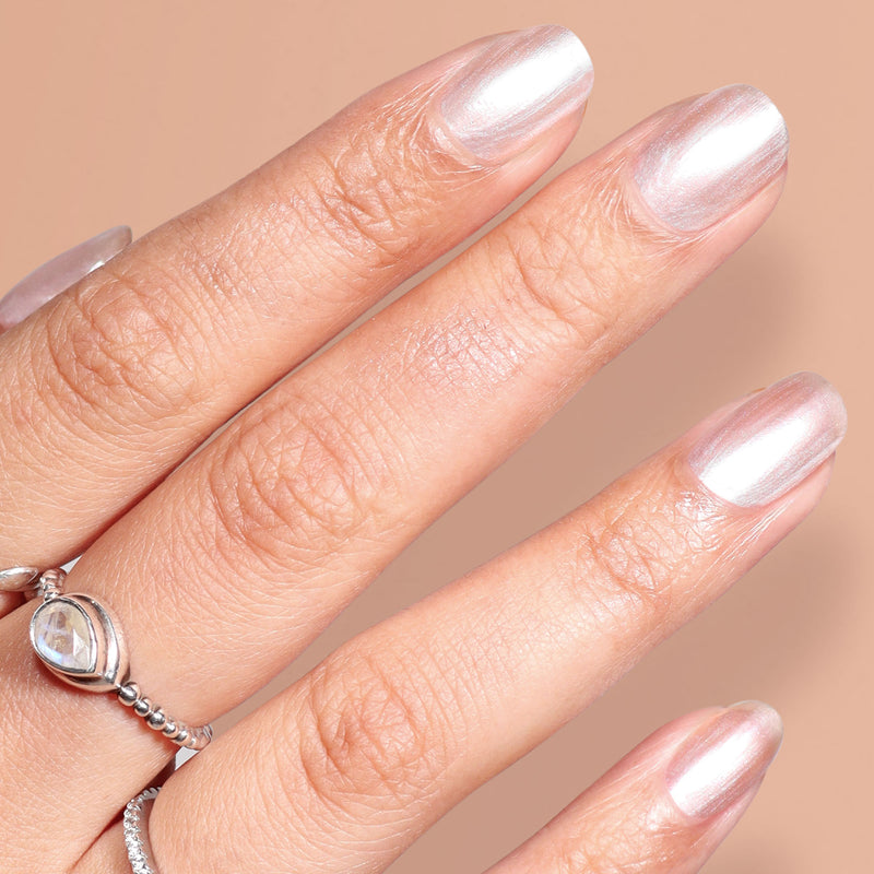 A close-up of a hand with oval-shaped nails painted in LONDONTOWN’s Cold Foam, a shimmery whipped pearl white polish, and wearing a silver ring with a clear gemstone.