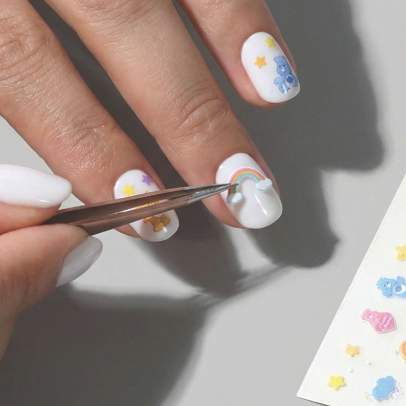 A hand uses tweezers to apply a Care Bears Nail Decal from LONDONTOWN onto a white-painted nail, surrounded by stars and cartoon animals for playful, creative nail art made easy.
