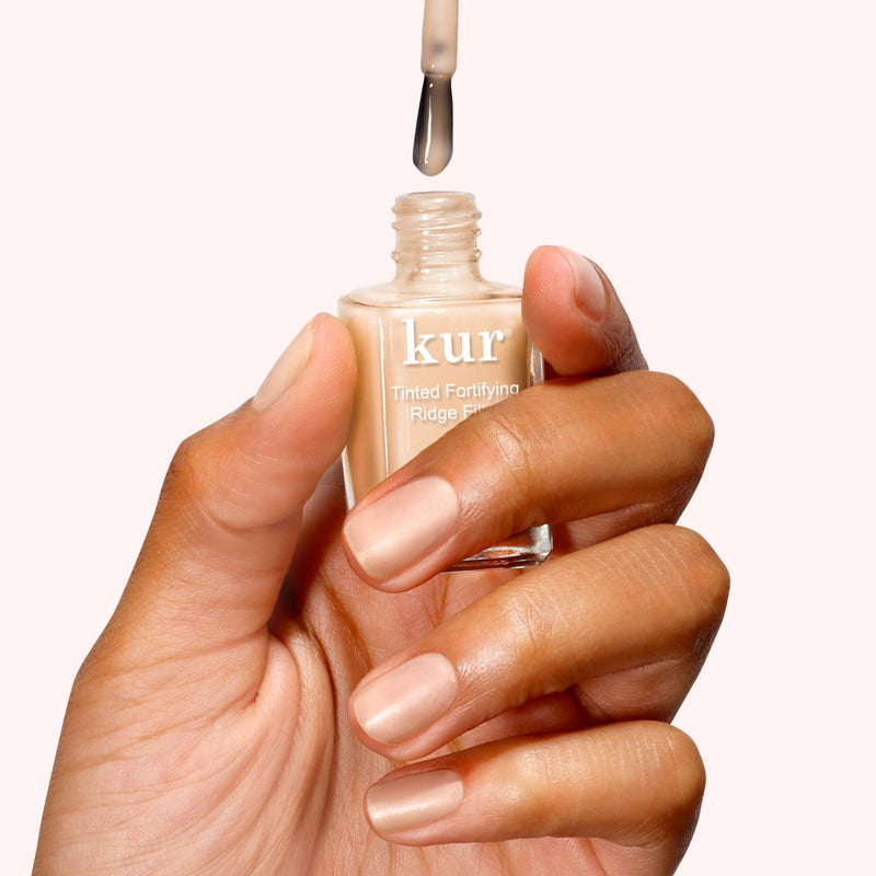 A hand delicately applies LONDONTOWN's Tinted Fortifying Ridge Filler in Apricot Beige to natural nails, subtly enhancing their color against a simple backdrop.