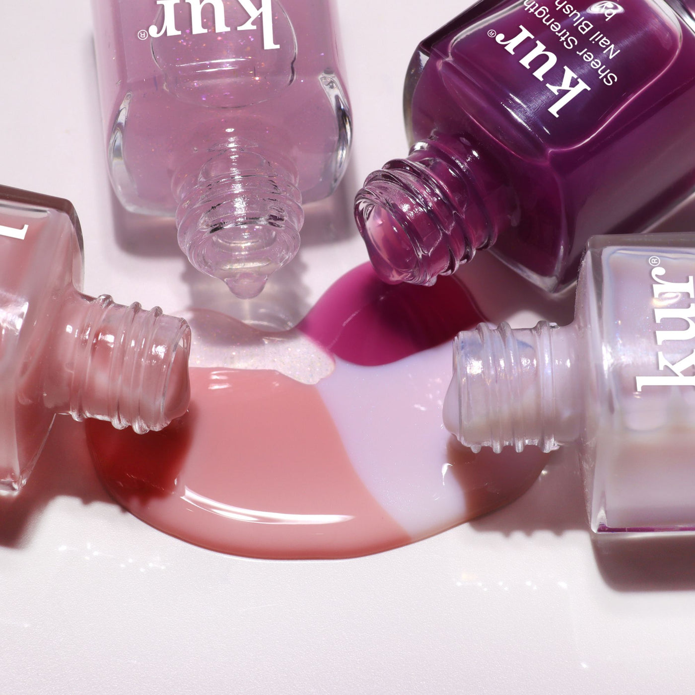 Three open LONDONTOWN One-Step Hero Minis nail polish bottles in pink and purple shades sit on a white surface, with their spilled colors blending together.