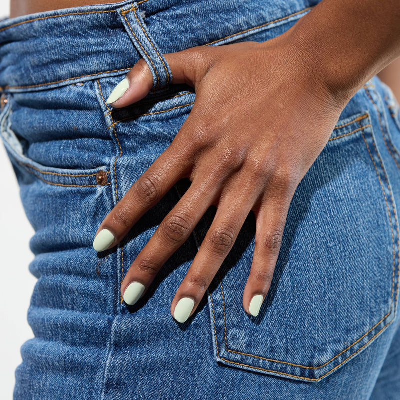 A hand with LONDONTOWN's "Lichen" nail polish, a pale green shade, rests on the hip of a person in blue denim jeans, evoking a calming vibe similar to white sage.