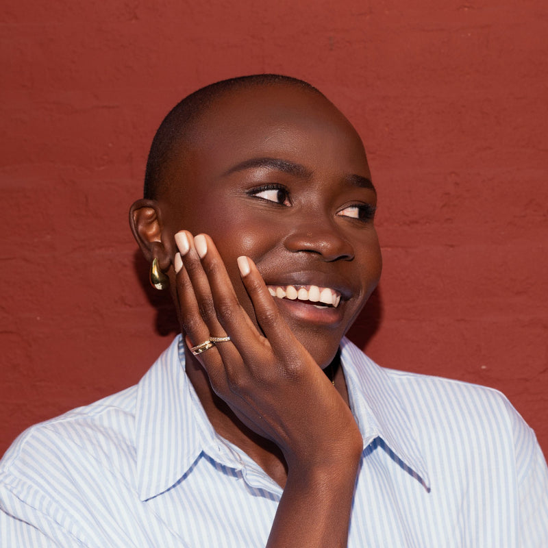 A person with dark skin and short hair smiles while touching their face, showcasing LONDONTOWN’s Moonlit Botanica Mini lakur trio, wearing a light blue striped shirt, gold hoop earrings, and a ring against a red brick wall.