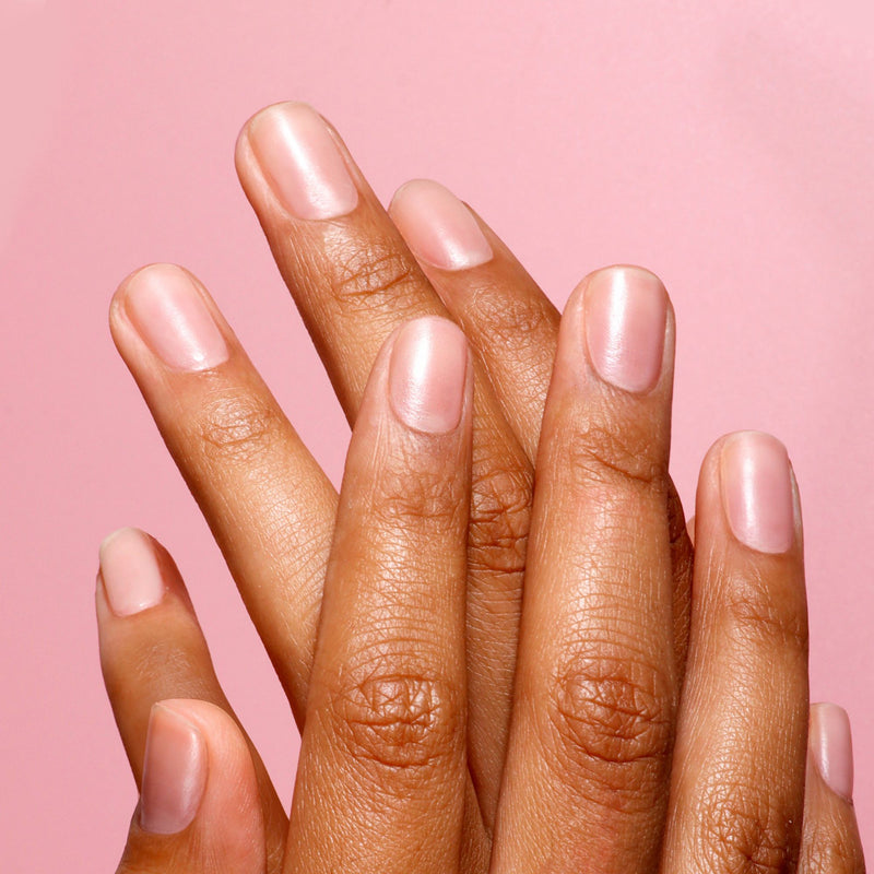 A close-up on a pink background features two hands with short, manicured nails treated with LONDONTOWN's Tinted Fortifying Ridge Filler in Petal Pink, highlighting smooth ridges and a subtle tint.