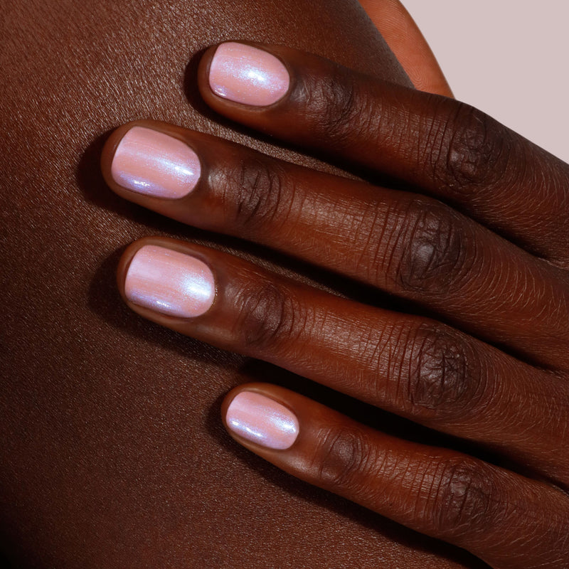 A hand with smooth, dark skin rests on a surface. The nails are neatly manicured and adorned with LONDONTOWN's Gel Genius Base Coat in shimmery light pink, delivering a chic look that's damage-free.