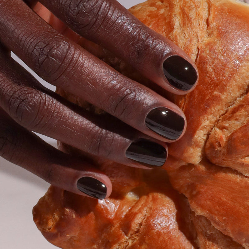 A hand adorned with LONDONTOWN’s Midnight Mocha nail polish holds a golden, flaky croissant next to a cup of rich espresso.
