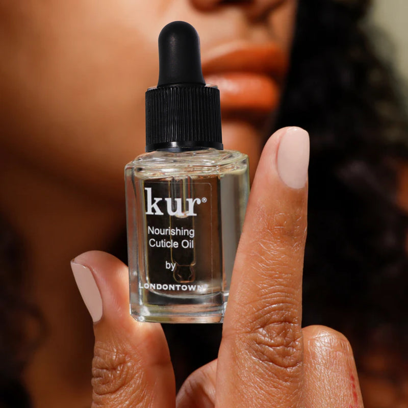 A hand with manicured nails holds a small glass bottle of LONDONTOWN Nourishing Cuticle Oil with a dropper cap, in front of a blurred face—ideal for enhancing cuticle care and supporting nail health.