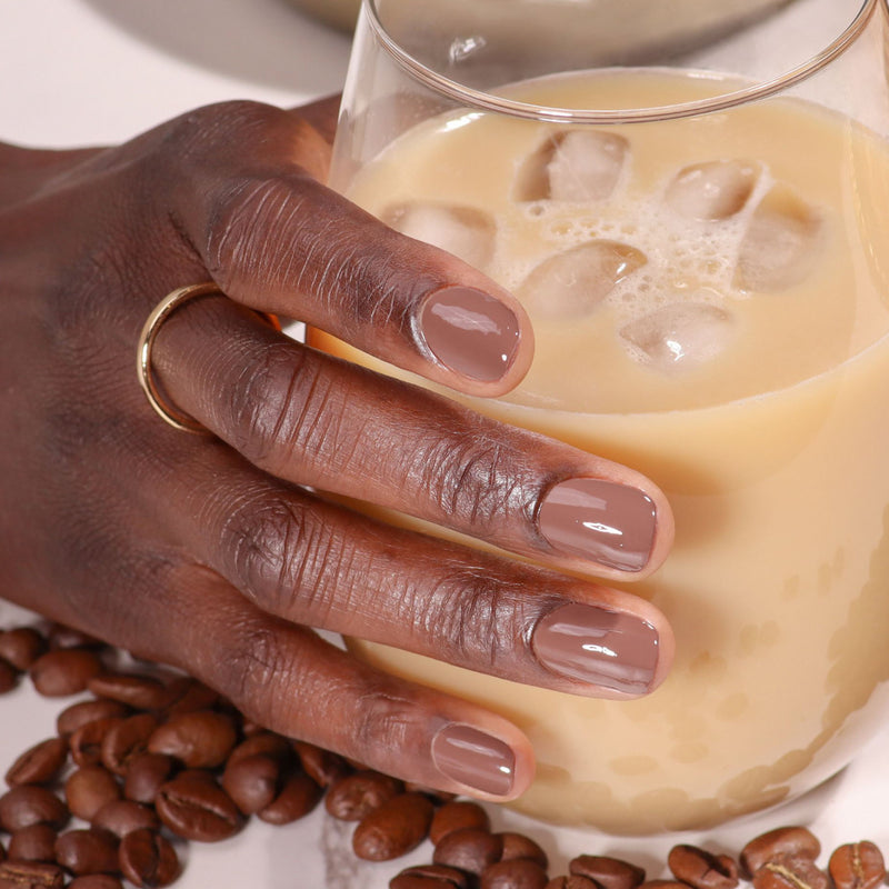 A hand with manicured Dirty Chai by LONDONTOWN nails holds a glass of iced coffee, with coffee beans scattered on the table.