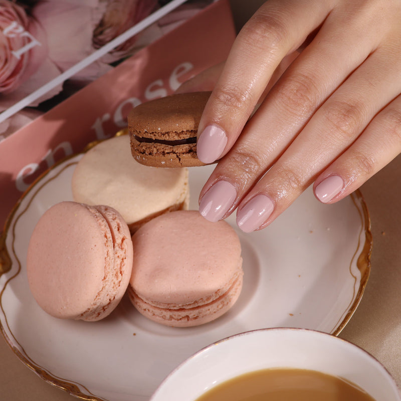 A hand with perfectly manicured, damage-free nails, thanks to LONDONTOWN's Gel Genius Base Coat, holds a chocolate macaron over a plate of three. In the background are a teacup and book offering perfect relaxation.