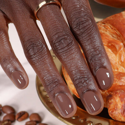 A hand with short, glossy nails in LONDONTOWN’s Dirty Chai shade rests on a gold plate beside a croissant and coffee beans.