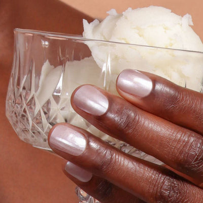 A hand with LONDONTOWN Cold Foam light pink metallic nail polish holds a glass dessert bowl with two scoops of white sorbet, capturing the smooth elegance of whipped pearl cream.