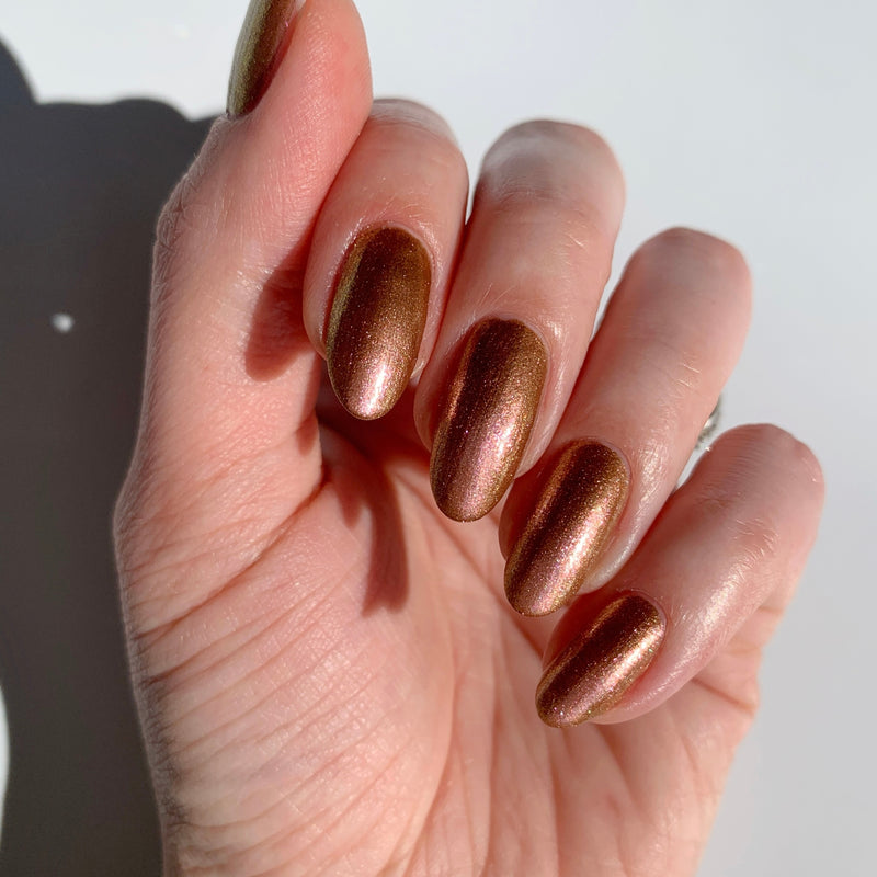 A hand with long, almond-shaped nails painted in LONDONTOWN's "The Runway Edit" metallic bronze polish is shown from above against a light background.