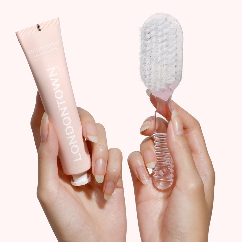 One hand holds a tube of LONDONTOWN's Mani Bright Nail Scrub, while the other grasps a clear nail brush, poised to brighten discolored nails against a light background.