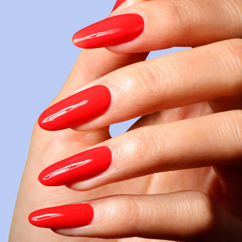 A close-up of a hand with long, almond-shaped nails painted in LONDONTOWN’s Maraschino Cherry, set against a light blue background.