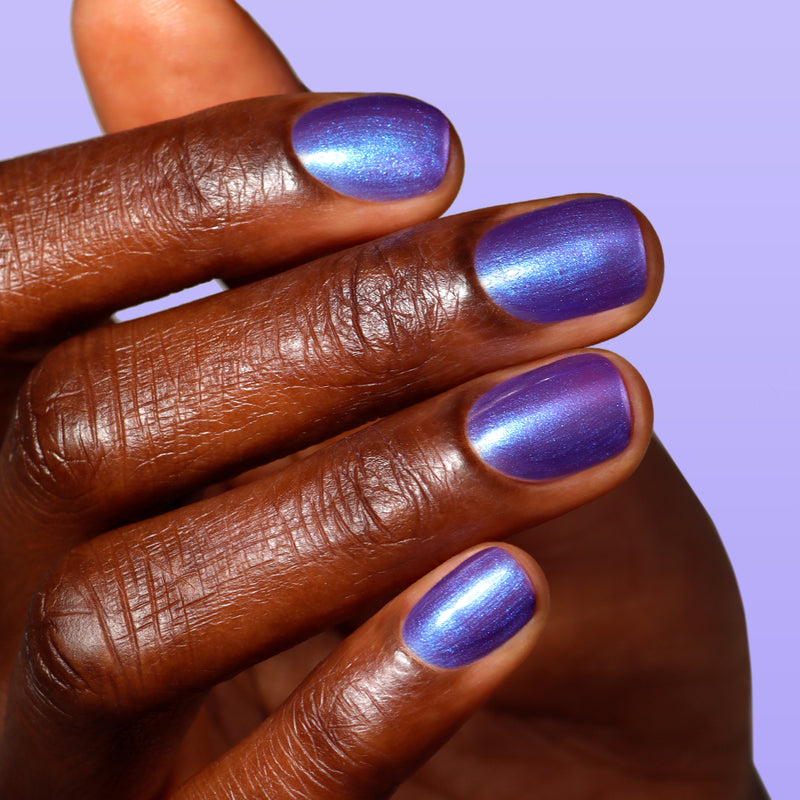 A hand with dark skin displays neatly manicured nails painted with LONDONTOWN’s Wildberry Freeze, a metallic blue-purple polish with a blue shimmer shift, shown against a light purple background.