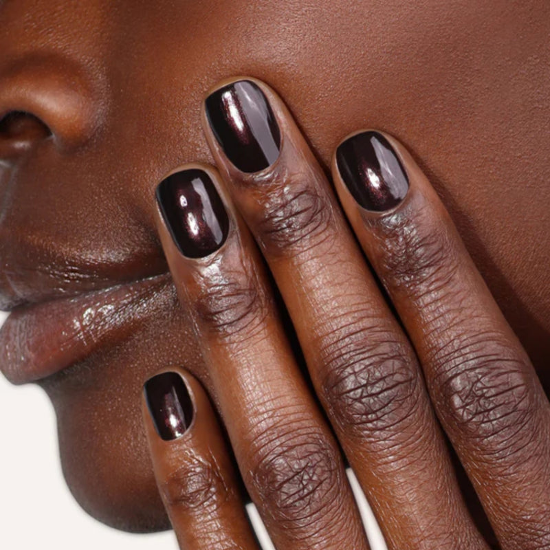 In the warm glow of candlelight, a person gently touches their cheek with fingers adorned in nails painted in LONDONTOWN's Spilled Wine, a dark metallic purple reminiscent of rich pinot noir.