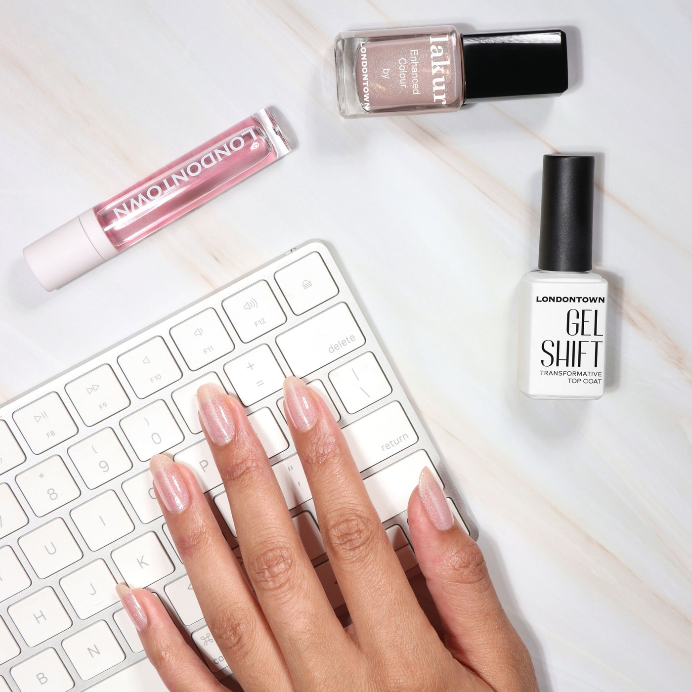 A hand with a gel manicure using LONDONTOWN GelShift Top Coat rests on a white keyboard, with polish bottles and a tube of lip gloss nearby on a white surface.