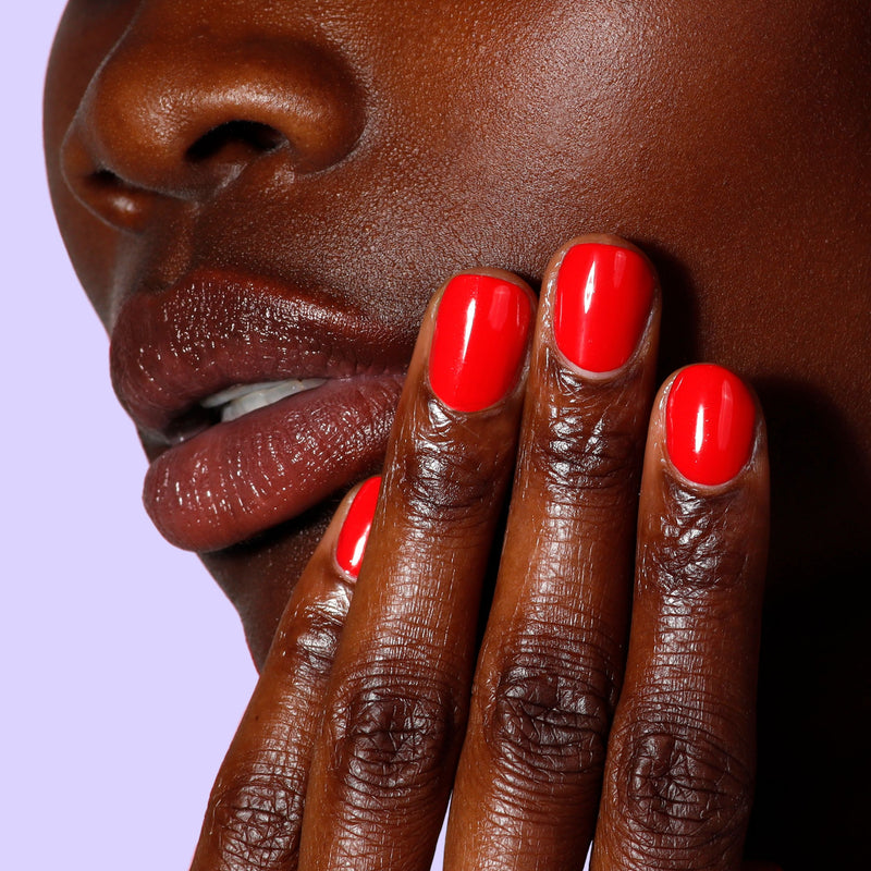 Close-up of a person with dark skin, lips in brown lipstick, and fingers painted with LONDONTOWN Maraschino Cherry—a glossy, bright red nail polish—against a light purple background.
