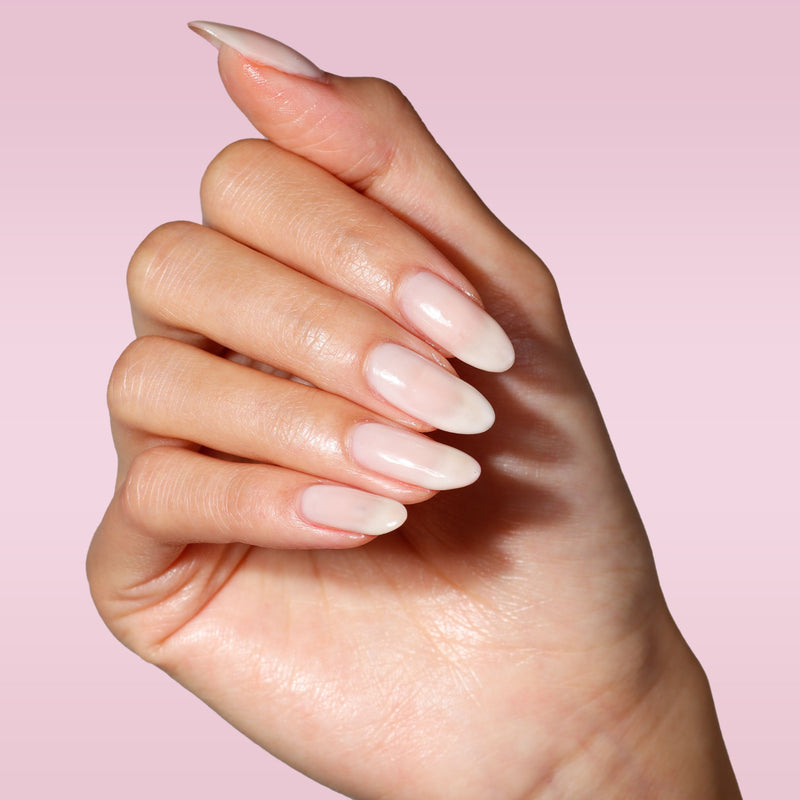 A hand with almond-shaped nails in LONDONTOWN's Double Scoop, a sheer pink, against a light pink gradient background.