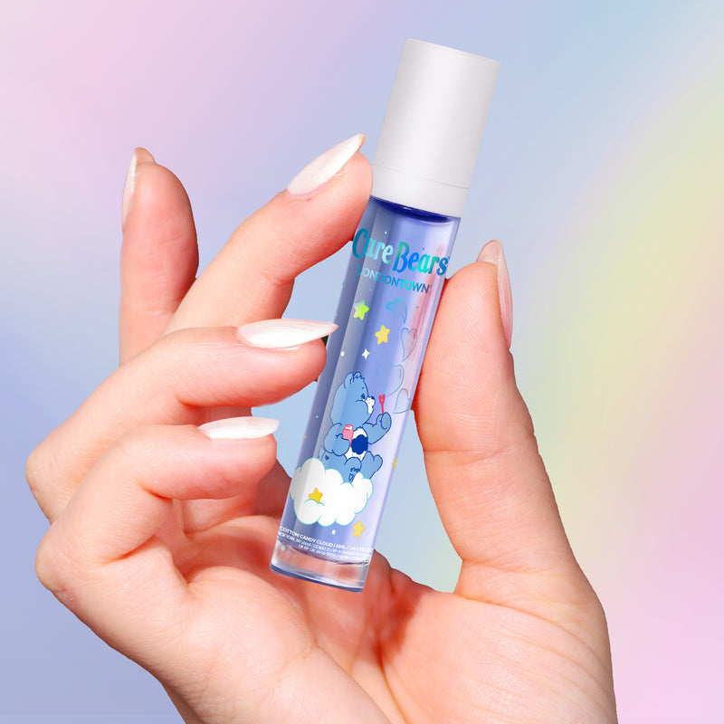 A hand with manicured nails holds LONDONTOWN’s Roll & Glow Cuticle Oil – Cotton Candy Cloud, featuring a Care Bears design, against a pastel gradient background.
