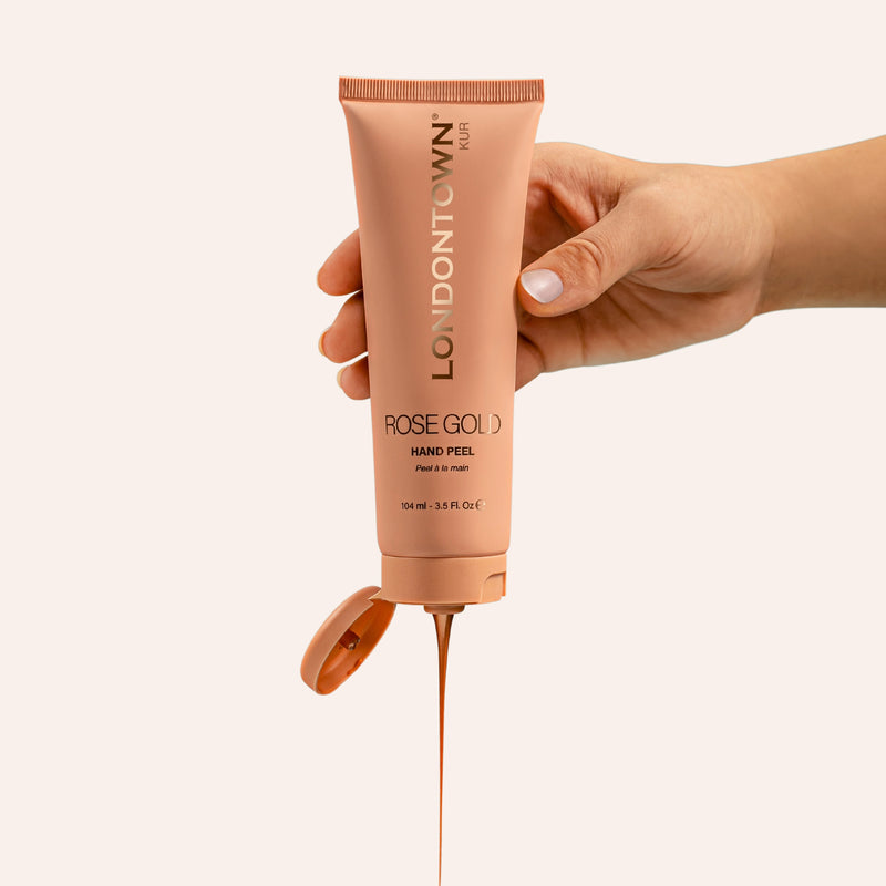 A hand holds a squeezed tube of LONDONTOWN Rose Gold Hand Peel against a neutral background, with the transformative mask dispensing from the nozzle, promising smoother, softer hands.