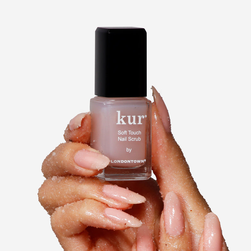 A hand with light pink nails holds a bottle of Soft Touch Nail Scrub by LONDONTOWN, showcasing exfoliated skin and visible granules. The bottle has a sleek black cap and the distinctive LONDONTOWN branding.