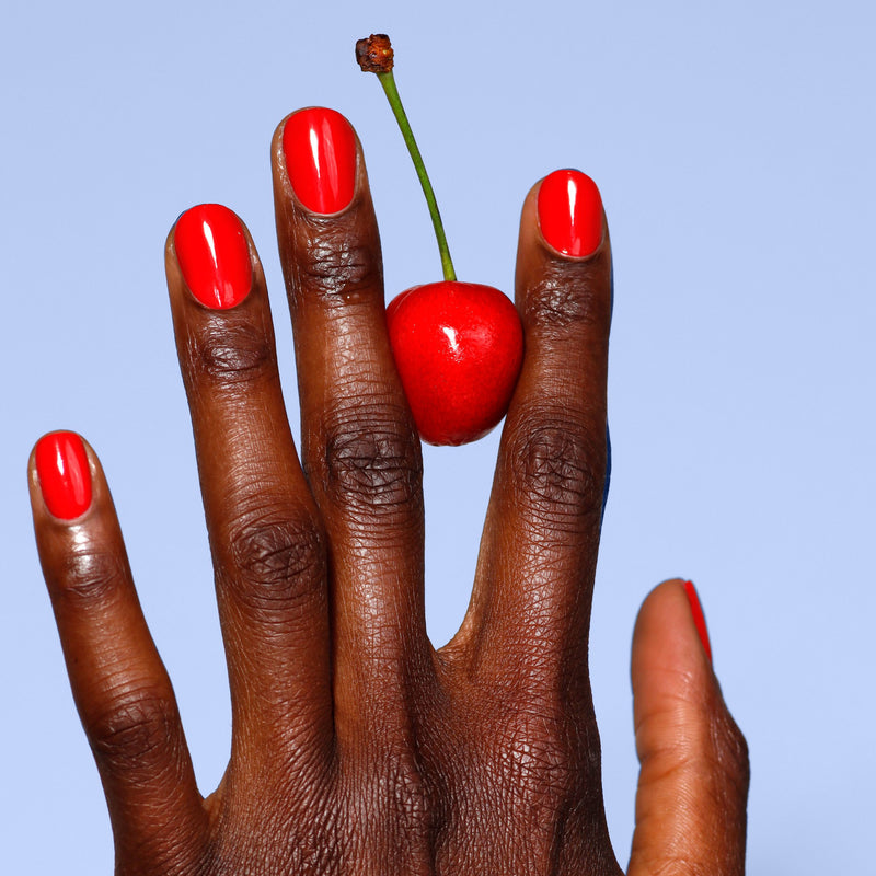 A hand with nails painted in LONDONTOWN's Maraschino Cherry shade delicately holds a cherry by its stem between two fingers against a light blue background.