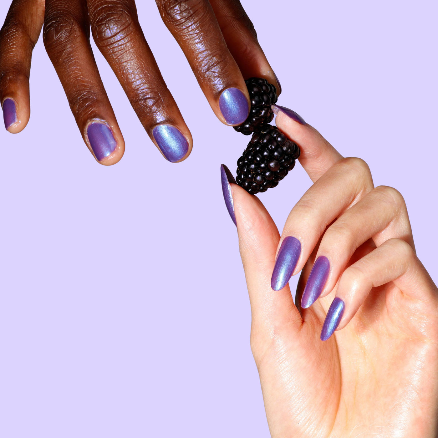 Two hands with LONDONTOWN's Wildberry Freeze, an iridescent purple polish with blue shimmer, hold blackberries against a light purple background.