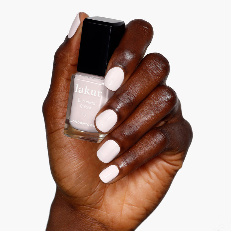 A hand with manicured powder pink nails holds a bottle of LONDONTOWN's Macaroon at Noon nail polish, exuding delicate charm against a pristine white background.