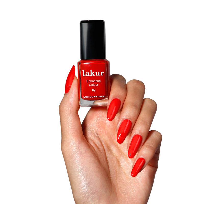 A hand with long, manicured nails painted bright red holds a bottle of LONDONTOWN Maraschino Cherry nail polish against a white background.