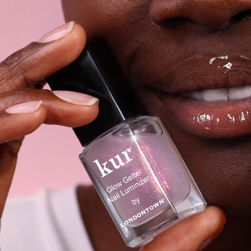 A person holds a bottle of LONDONTOWN Glow Getter Nail Luminizer near their glossy lips against a pink background, highlighting luminous nail care and an ethereal finish.
