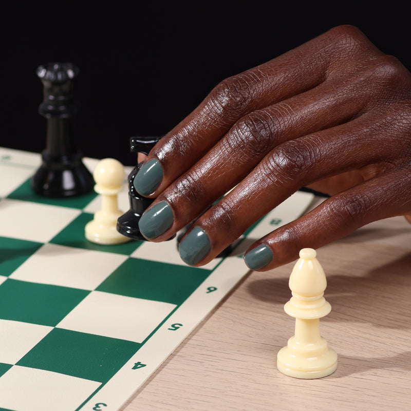 Someone with dark skin, wearing LONDONTOWN's Velvet Drapes olive green polish, moves a black bishop on a chessboard surrounded by white and black pawns.