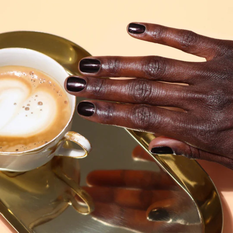 A hand with dark polished nails from LONDONTOWN rests on a mirrored surface by a cup of cappuccino and a glass of Spilled Wine on a gold tray.