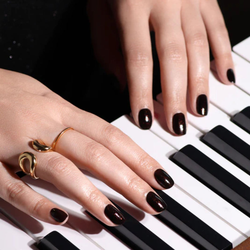 Close-up of hands adorned with LONDONTOWN's Around the Fireplace deep brown nail polish and copper-flecked rings playing a piano keyboard.