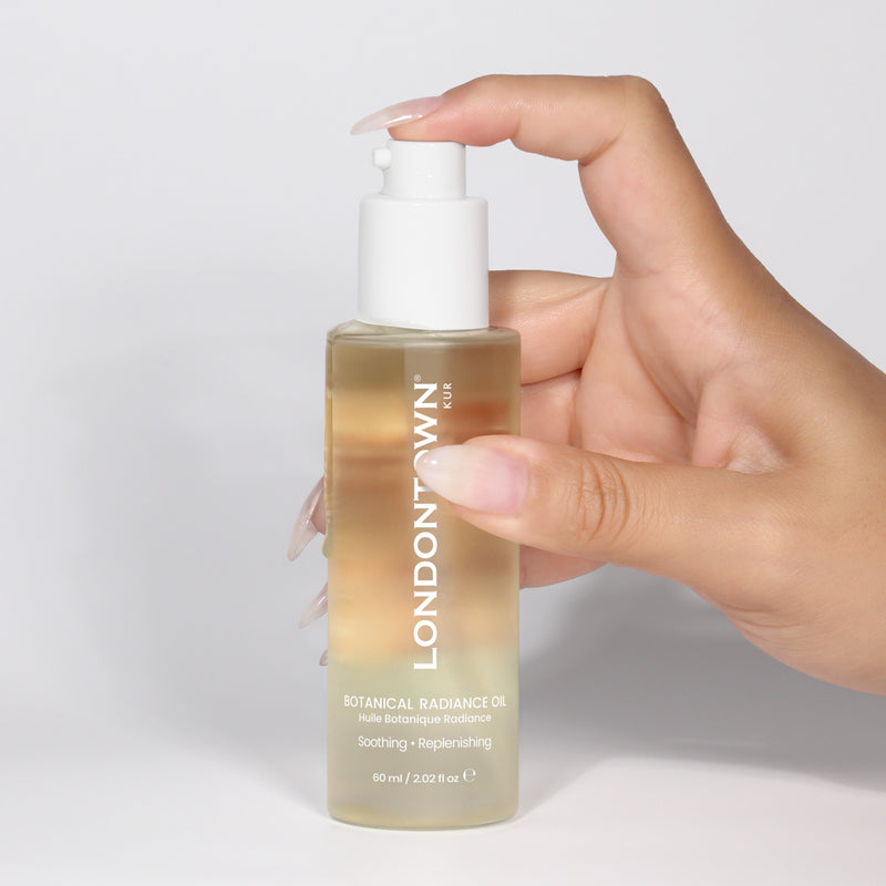 A hand holds a clear bottle of LONDONTOWN Botanical Radiance Oil, featuring a white pump dispenser, shown against a plain white background.