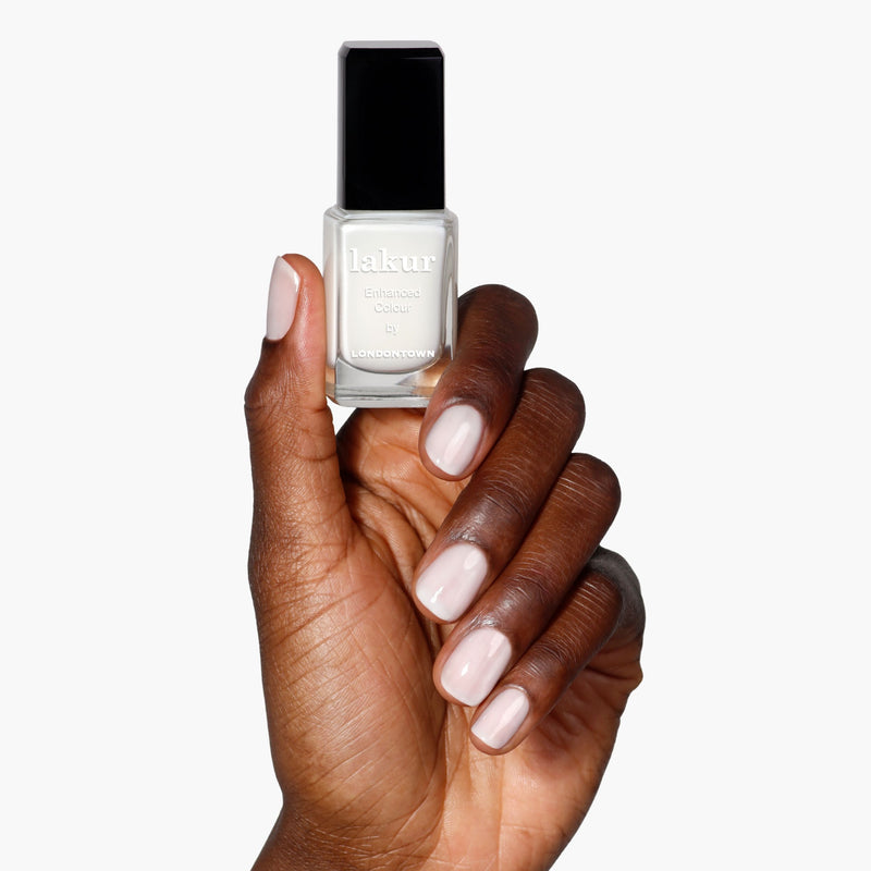 A hand with light pink manicured nails holds a bottle of LONDONTOWN Double Scoop, a creamy light grey nail polish, against a white background.