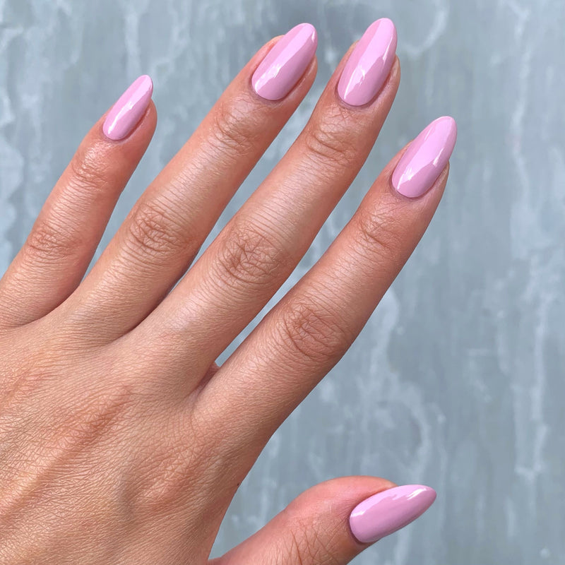 Against a gray textured background, a hand displays long, almond-shaped nails painted in LONDONTOWN's glossy Berry Nude, reminiscent of the soft allure of whipped cream.