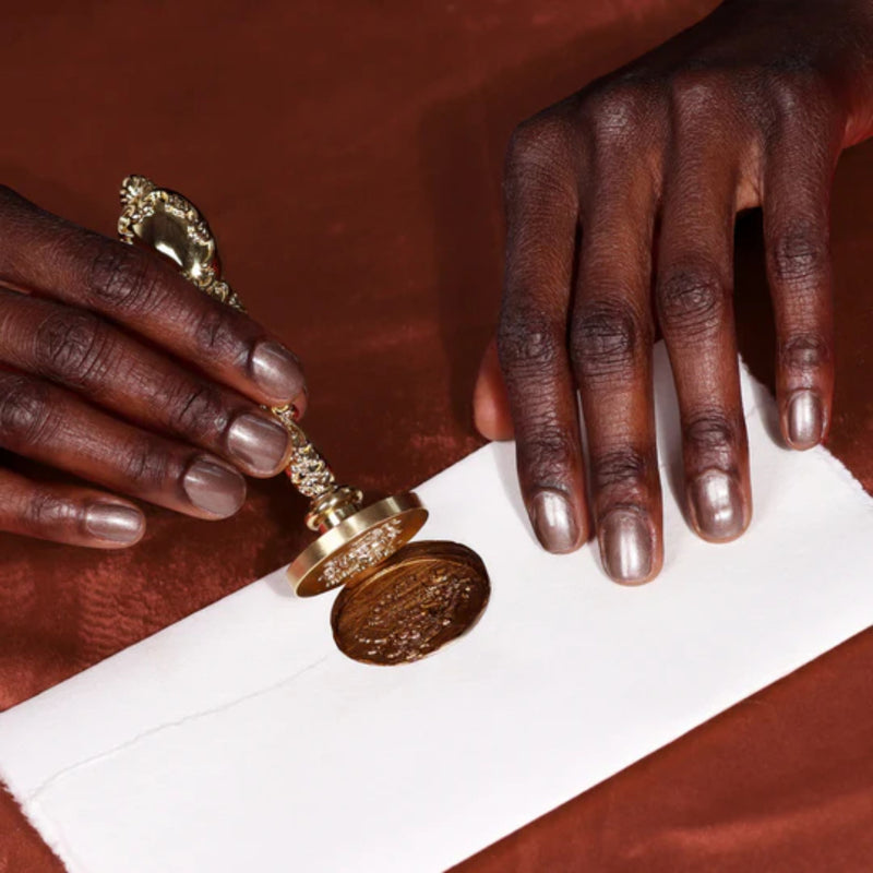 Hands using a Nightcap antique wax seal stamp by LONDONTOWN to seal an envelope on a bronze-toned surface.