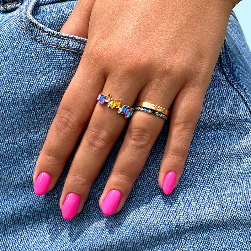A hand flaunts LONDONTOWN's bright pink vegan nail polish, Berry Punch, on a denim background. The look is completed with a gold band and a colorful gem-studded ring for added flair.