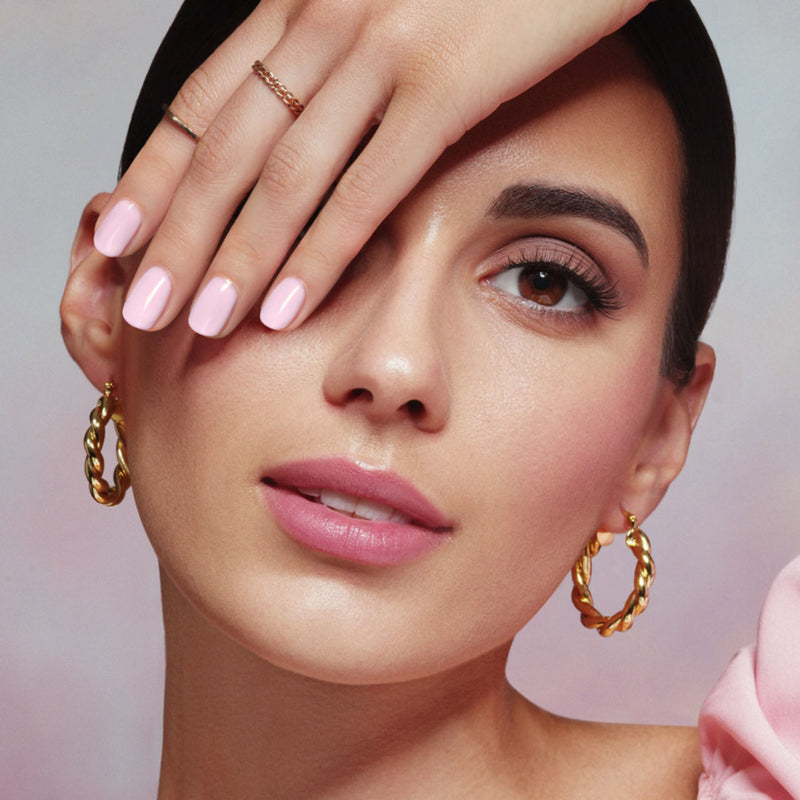 Sporting LONDONTOWN's Waterloo Sunset rose petal pink nail polish, a person partially covers their face with their hand, adorned with gold twisted hoop earrings and a slicked-back hairstyle, exuding cruelty-free elegance.