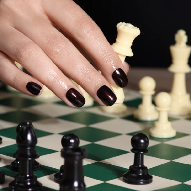 A hand with Spilled Wine, LONDONTOWN's dark nail polish, glimmers in candlelight as it moves a white rook on a chessboard amid black and white pieces.