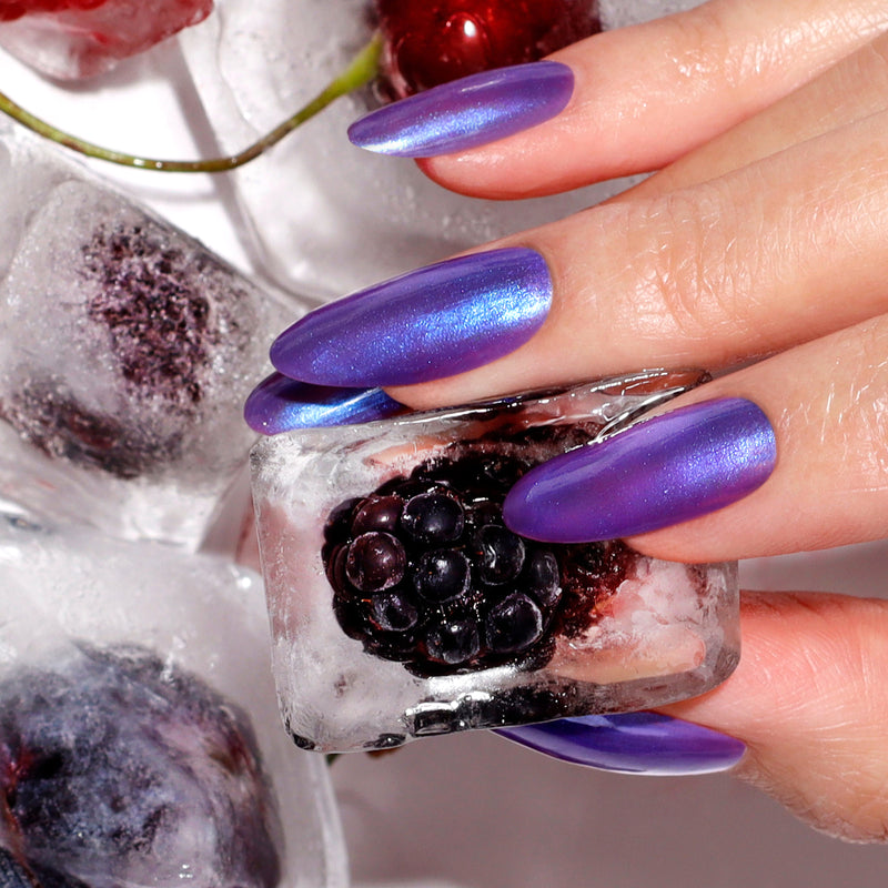 A hand with metallic purple-blue shimmer nails, painted with LONDONTOWN's Wildberry Freeze, holds an ice cube containing a blackberry; more berry-filled ice cubes appear in the background.